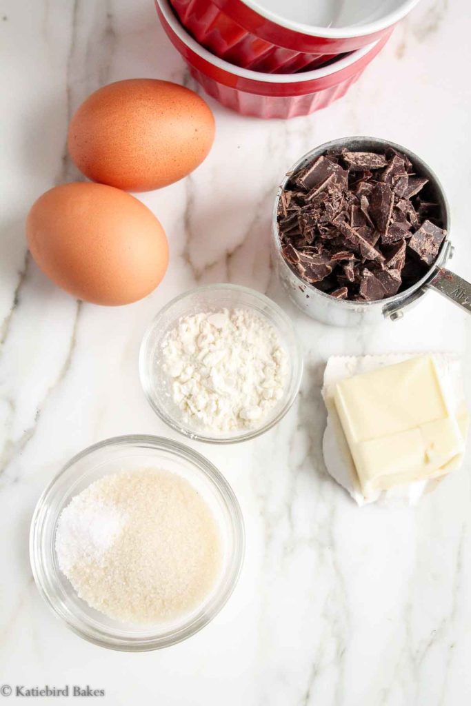 Ingredients on a white marble surface: 2 ramekins, 2 large eggs, chopped dark chocolate, flour, butter, sugar