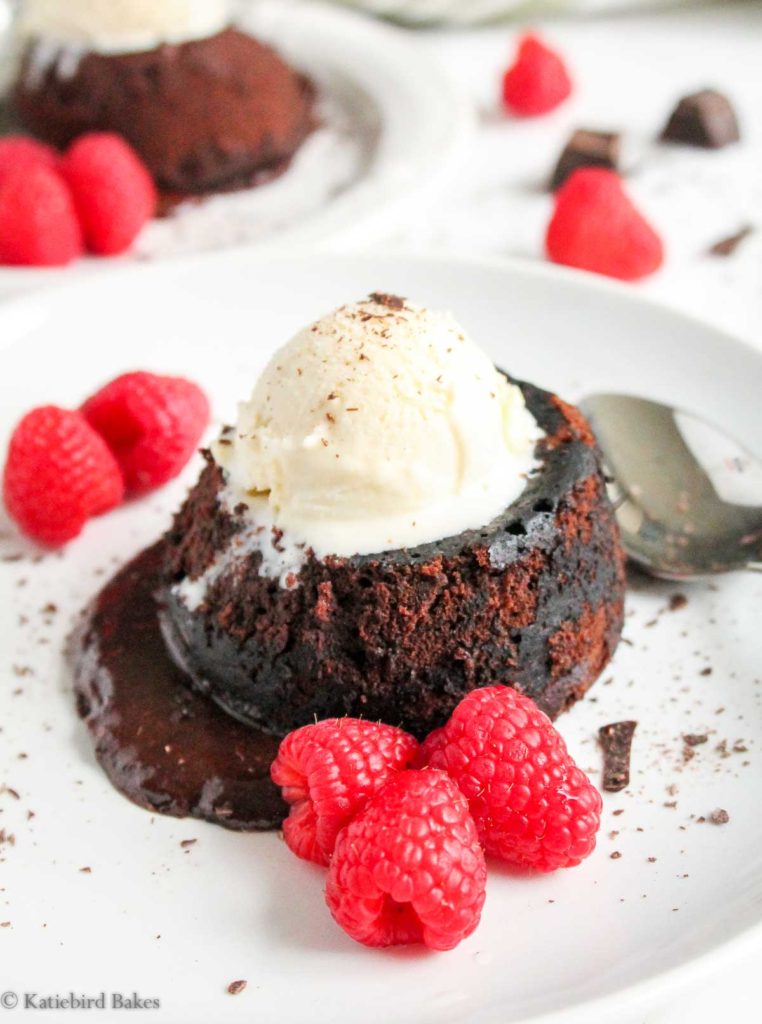 A head on view of one of the Molten Chocolate Lava Cakes for Two on a plate, with vanilla ice cream on top, surrounded by raspberries with chocolate oozing out of the cake