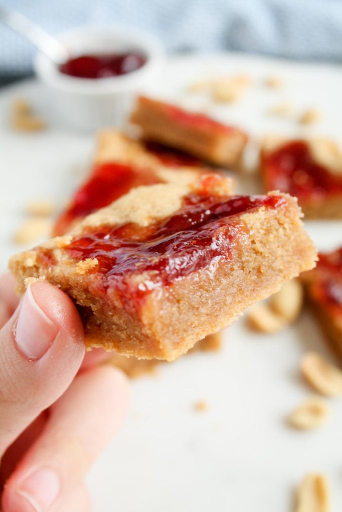 A hand holding one finished cookie bar with strawberry jam visibly swirled on the top