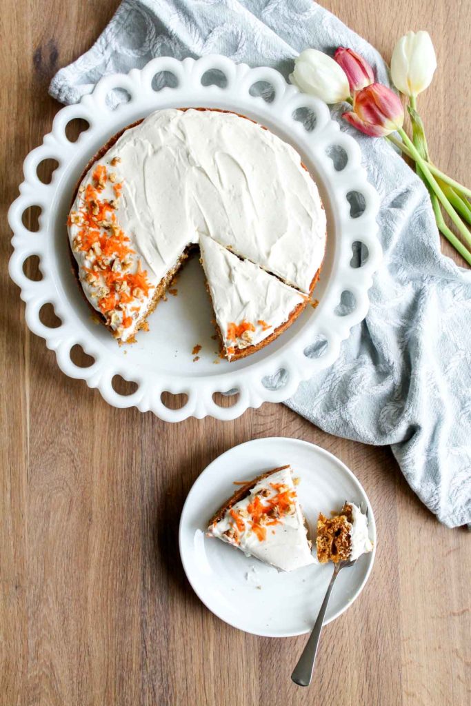 Overhead view of a slice of Single Layer Carrot Cake with Cream Cheese Frosting on a plate on a wood background, with the rest of the cake on a serving platter in the background.