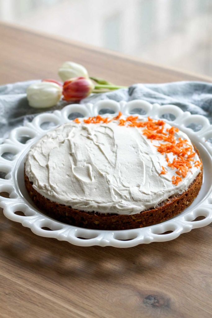Frosted cake on a cake plate and wood surface, with decorative grated carrots on top