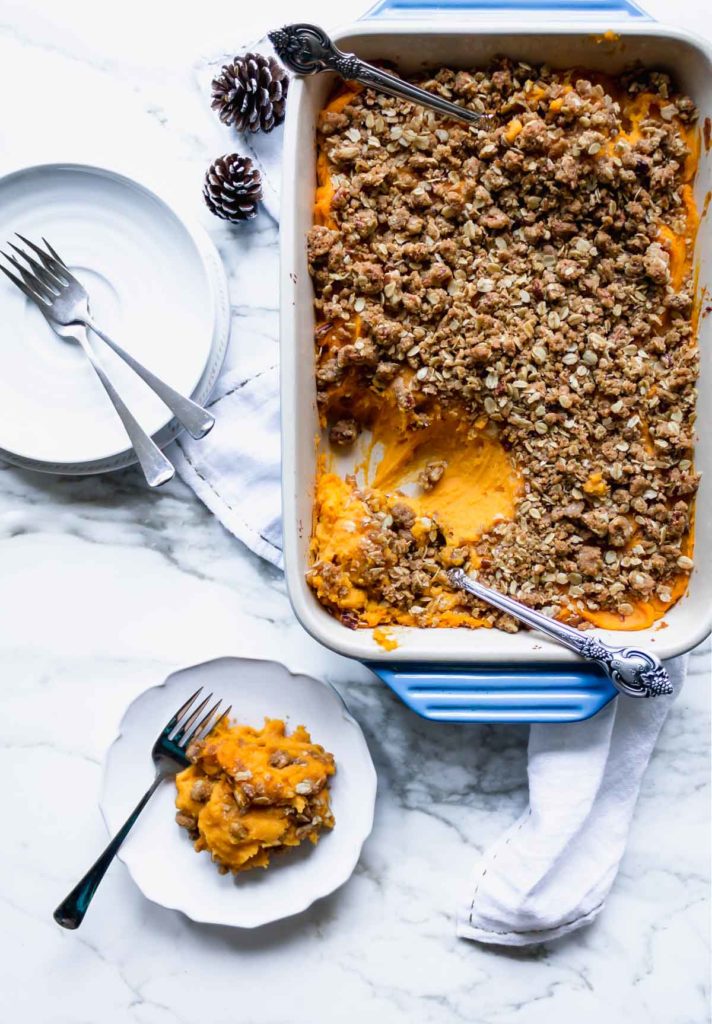 a serving of healthier sweet potato casserole with pecan topping on a plate, with the baking dish in the background