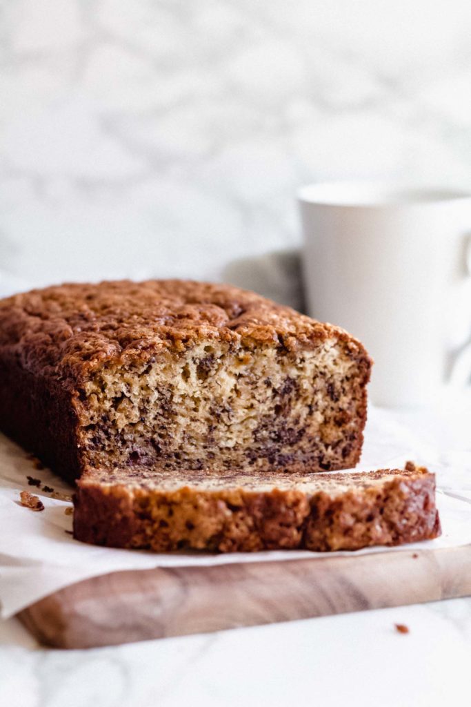 one slice of this easy banana bread recipe next to the rest of the loaf, sitting on a cutting board with a coffee cup in the background