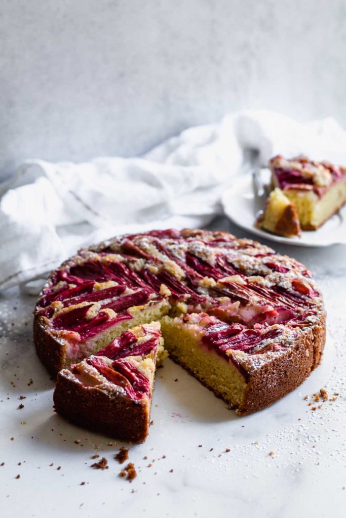 45-degree view of the rhubarb coffee cake with a couple slices cut out, one on a plate next to the cake, on a white background with a white tea towel