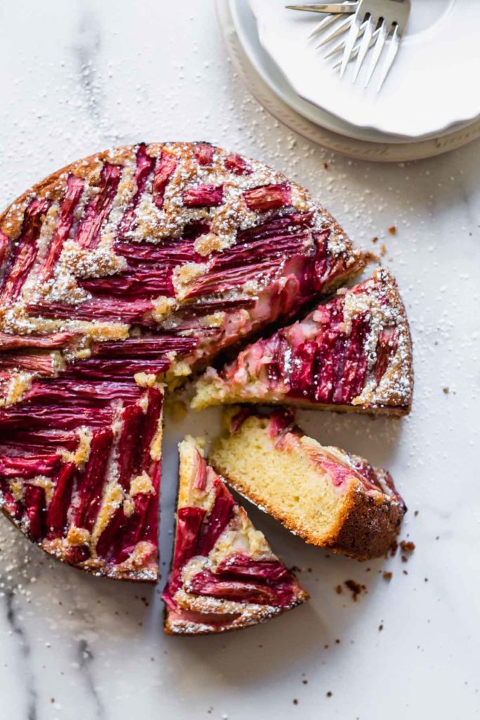 overhead view of slices of rhubarb coffee cake arranged on a white marble background with plates and forks off to the side
