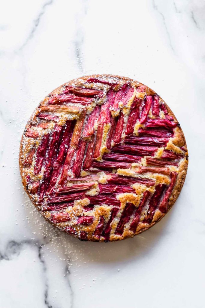 overhead view of baked Rhubarb Coffee Cake on a white marble surface, with visible chevron pattern of red rhubarb on top of the cake and powdered sugar sprinkled over top