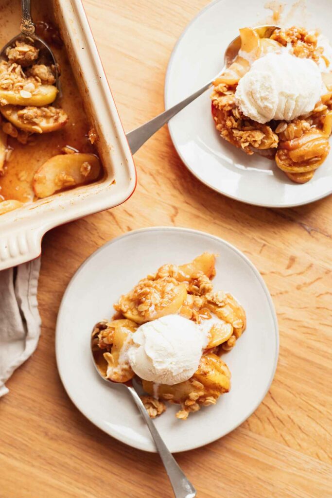 2 bowls of easy apple crisp with oat topping and vanilla ice cream on a table next to the baking dish with a serving spoon