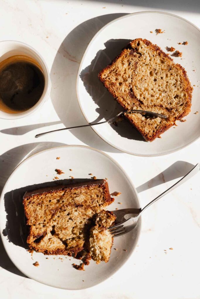 two slices of cinnamon swirl banana bread on white places with forks cutting into them and a half drunk cup of coffee on the side