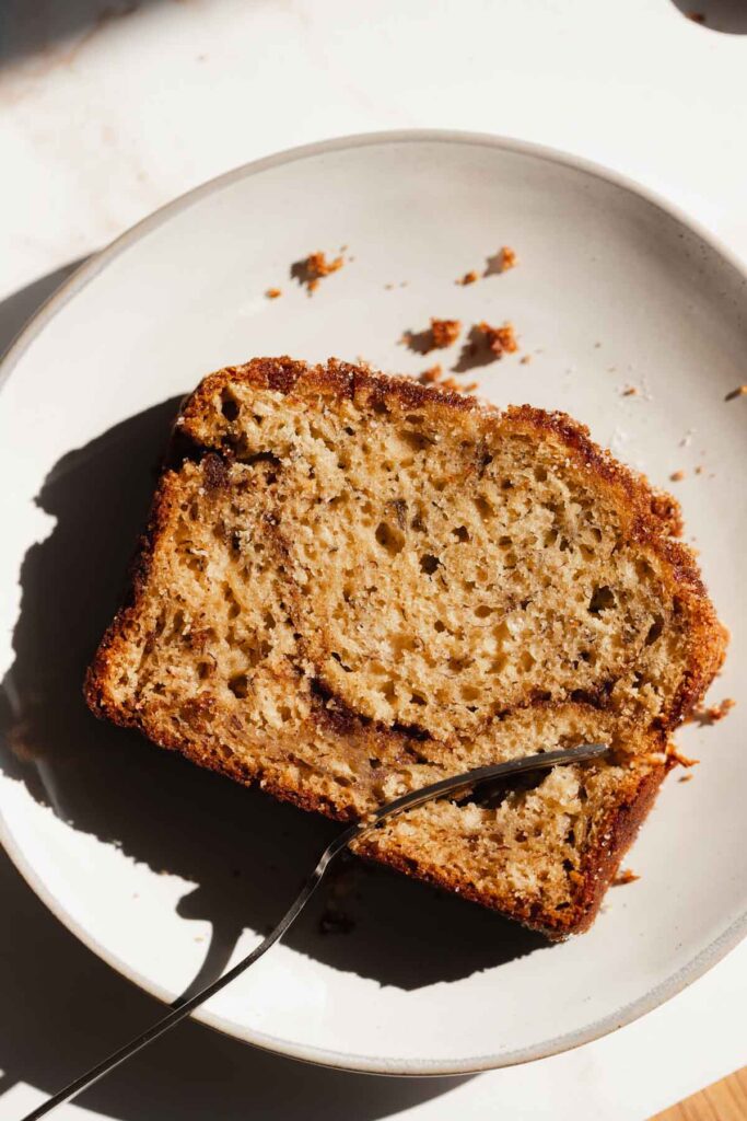 a slice of cinnamon swirl banana bread on a white plate with a fork cutting into it